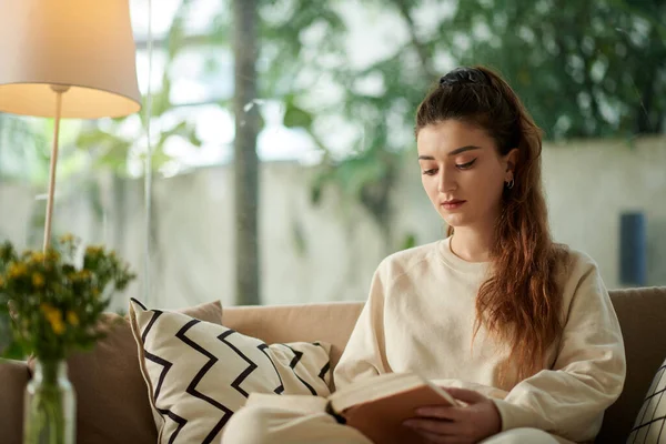Serious young woman reading good book when resting on sofa at home