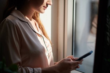 Smiling girl standing at window and texting friend