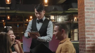 Restoranda çocuklu bir ailenin siparişini alırken zarif üniformalı Asyalı garsonun dijital tablet kullanmasını çek.