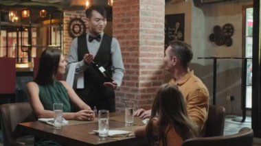 Klasik üniformalı bir garsonun masaya şişe getirmesi ve ailesine restoranda servis yaparken şarap eşleşmesi önermesi.