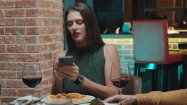 Güzel bir kadının restoranda masada otururken akıllı telefonuyla yemeklerin fotoğrafını çektiği orta boy bir fotoğraf.