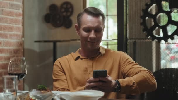 中等比例的白人男子坐在餐桌前 一边用智能手机一边吃饭一边和别人说话 — 图库视频影像