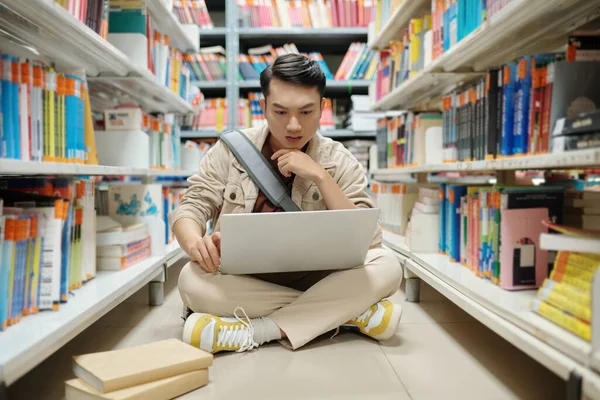 认真的大学生坐在图书馆的地板上 在笔记本电脑屏幕上阅读信息 — 图库照片