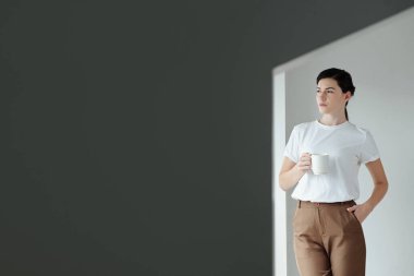 Düşünceli genç bir kadın minimalist tasarımla odada kahve içiyor.