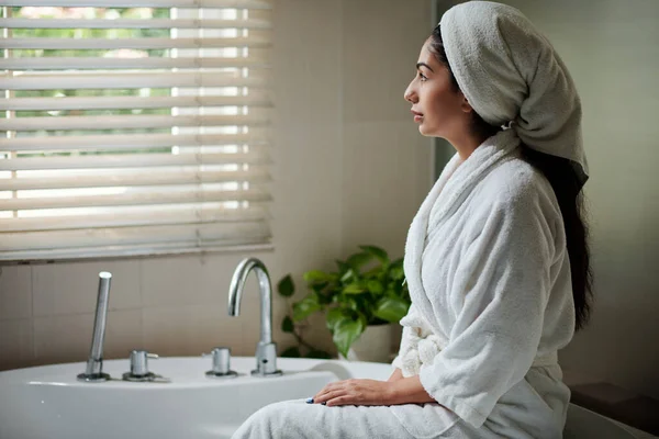 Young woman in bathrobe and towel on head sitting next to bathtub