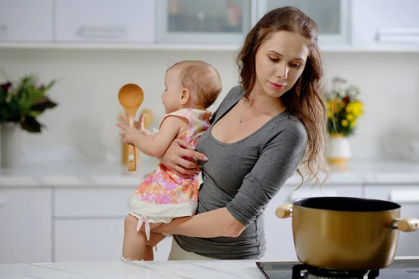 Young woman holding little daughter when cooking soup or pasta for dinner at home