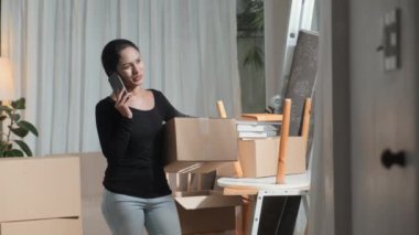 Paketlenmiş eşyaların arasında duran Asyalı kadın, elinde karton kutuyla yeni eve taşınırken cep telefonuyla konuşuyor.