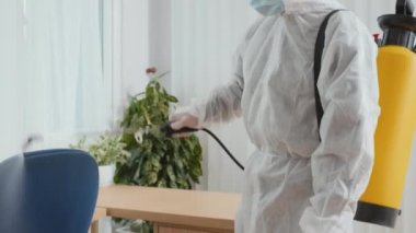 Koruyucu takım elbiseli temizlik işçisi ve yüz maskesi dezenfektanı müşterinin evini temizlerken bir galonluk mobilyanın üzerine sıkarken orta ölçekli bir fotoğraf.