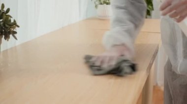 Koruyucu takım elbiseli temizlik görevlisinin ev masasını temizlerken dezenfekte spreyi ve bez kullanarak yakın plan çekimleri.