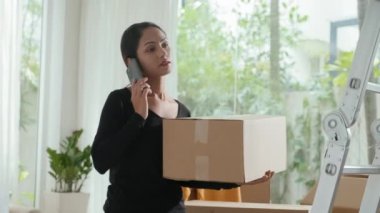 Genç esmer kadının paketlenmiş kutusunu tutarken ve taşınmaya hazırlanırken telefon ederken orta boy fotoğrafı.