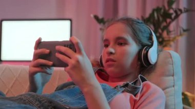 Neşeli Kafkasyalı gen z kızı, karanlık bir odada, neon pembe ışıklandırmayla kanepede uzanırken akıllı telefonuyla oyun oynuyor.