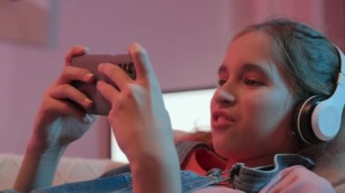 Kablosuz kulaklıklı genç oyuncu kız odasındaki kanepede dinlenirken akıllı telefonuyla oynuyor.