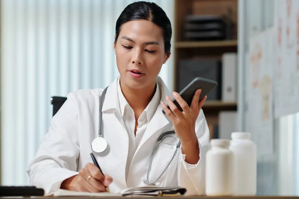 研究室のコートを着た若い女性臨床医は 医療事務所に座っている間 スマートフォンで電話をかけてメモや処方をしています — ストック写真