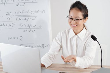 Evden uzaktan öğretmenlik yapan gülümseyen genç kadın, arka planda matematik formülleri olan beyaz tahta.