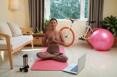 Evde yoga minderinde meditasyon yaparken dizüstü bilgisayar başında ders izleyen gülümseyen genç bir kadın.