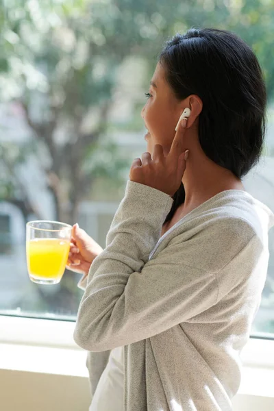 积极的年轻女性在喝果汁时戴着耳塞 透过窗户看去 给朋友或同事打电话 — 图库照片