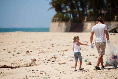 Baba ve kızı kirli sahilde yürüyor ve Dünya gününde plastik atık topluyor.