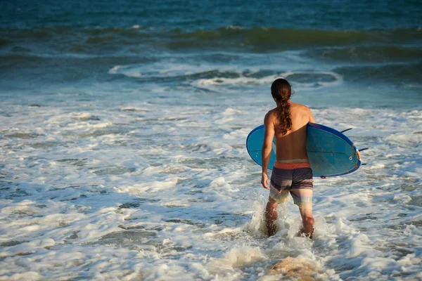 長い髪のサーフボードを運ぶと海に歩いて若いサーファー 背中からの眺め — ストック写真