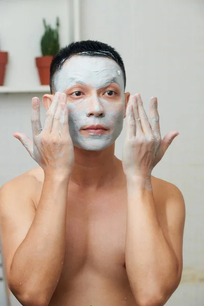 Shirtless Jovem Aplicando Desintoxicante Máscara Barro Branco Rosto Quando Espelho — Fotografia de Stock