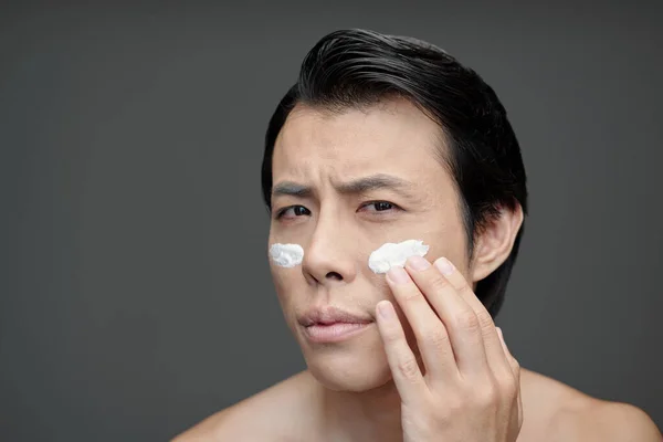 Frowning man applying moisturizer or sunscreen on face