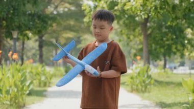 Mutlu küçük Asyalı çocuk, yaz günü parkta oynarken kameraya doğru koşuyor ve oyuncak uçak fırlatıyor.