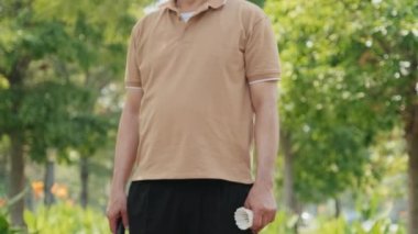 Mutlu, olgun Asyalı bir adamın yaz günü parkta elinde badminton raketi ve mekikle dikilip yeşil yapraklara karşı gülümseyerek poz verişini çek.