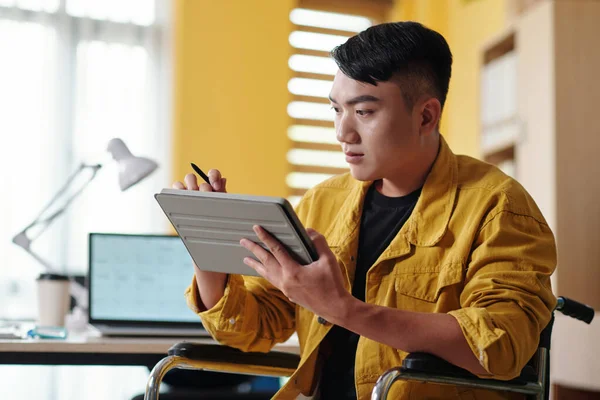 Young man with disability working in office of company, reading document on tablet
