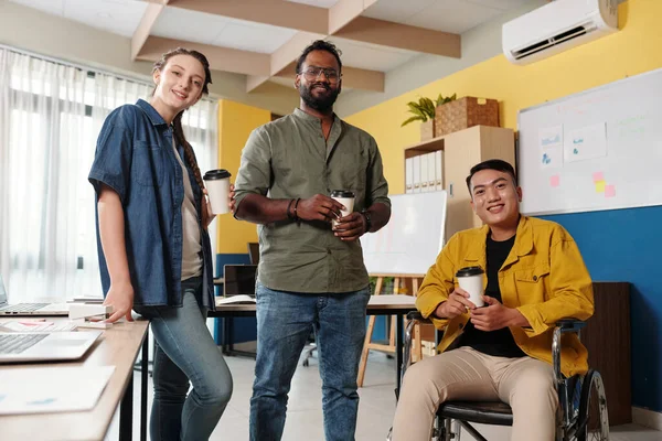 Positive diverse business team standing in open space office with take-out coffee in hands