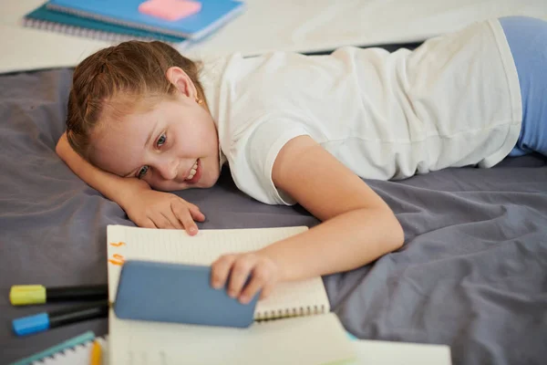 微笑的女孩躺在床上 在智能手机上观看有家庭作业的娱乐性视频 — 图库照片