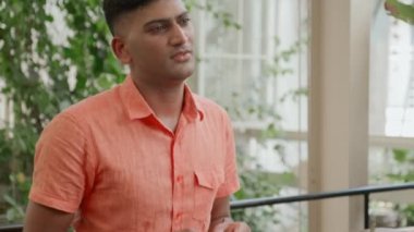 Kafede yeşil bitkilere karşı oturan, kahve içen ve iş arkadaşı veya arkadaşıyla konuşan genç Hintli bir adam.