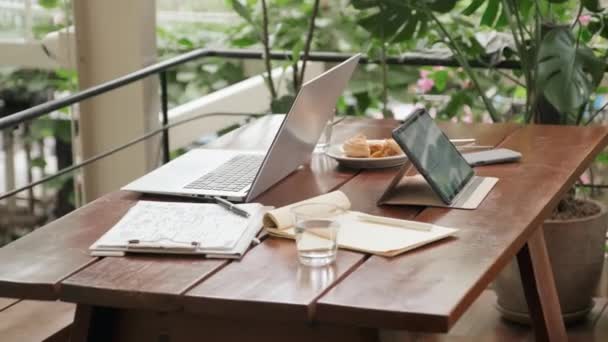 有数码平板电脑 笔记本电脑 商业文件和水杯的中景餐桌 咖啡馆里装饰着绿色植物 没有人 — 图库视频影像