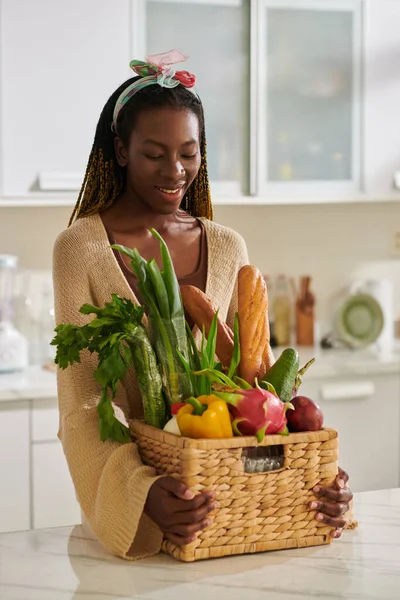 Portrait of smiling Black young woman putting basket of fresh groceries on kitchen counter