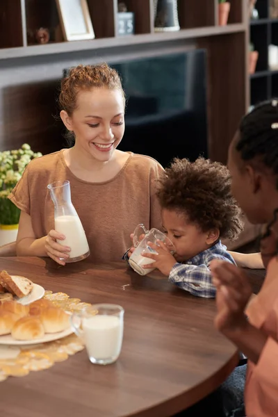 快乐的年轻母亲看着她的儿子在厨房桌上喝着一杯牛奶 — 图库照片