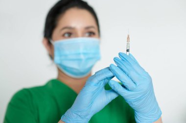 Şırıngayı aşıyla kontrol ederken eldiven ve tıbbi maske takan bir hemşire.