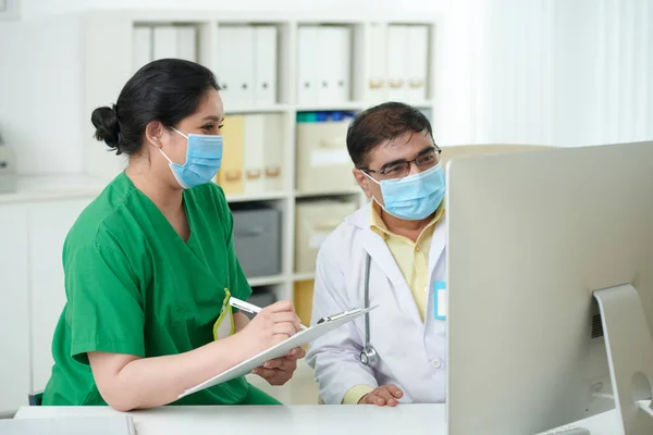 在电脑屏幕上讨论病人胸部X光时 戴防护面罩的医务工作者在文件中填入资料 — 图库照片