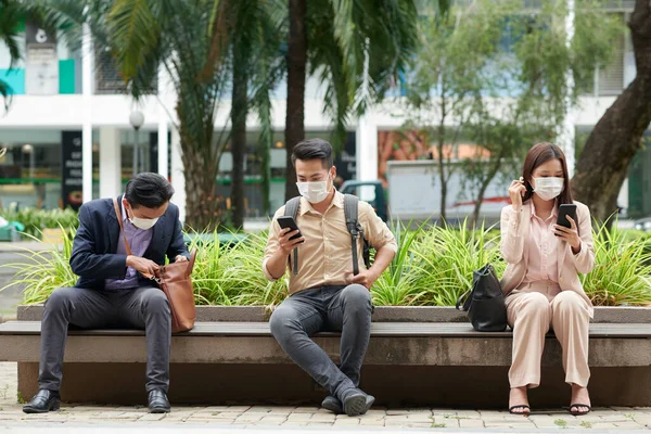 在午休时间 戴着防护面具的商人们坐在长椅上休息 听音乐 在社交媒体上看新闻 — 图库照片