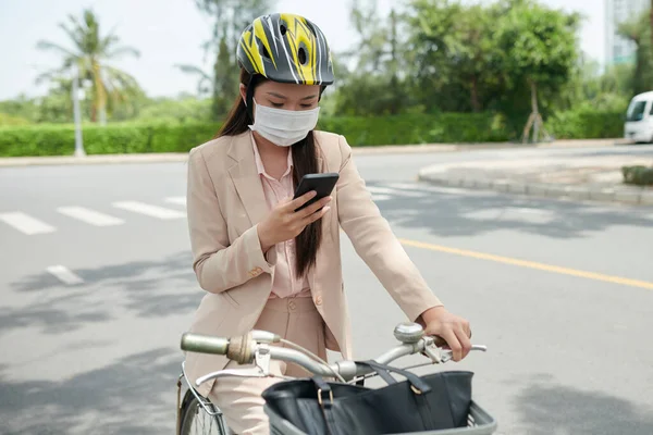 Young businesswoman in protective mask sitting on bicycle and checking notifications on smartphone