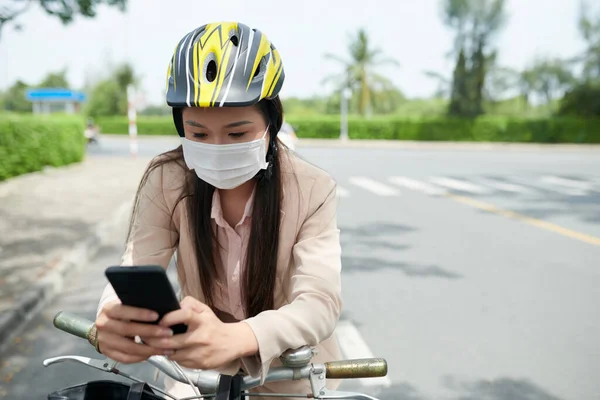 戴着防护头盔和医疗面罩的年轻女子骑自行车 用智能手机回复短信 — 图库照片