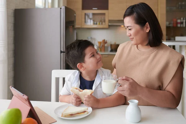 幸せな母親は朝食のために息子にミルクとハムとチーズサンドイッチのカップを与える — ストック写真