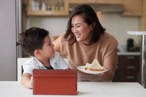 笑顔母もたらすプレートにサンドイッチで小さな息子に見教育ビデオのデジタルタブレット — ストック写真