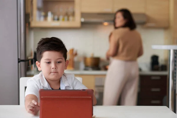 重大な男の子ビデオを見ますタブレットコンピュータ上で彼のお母さん料理夕食でバックグラウンド — ストック写真