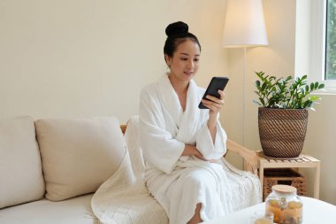 Bornozlu genç Vietnamlı olumlu bir kadın spa prosedüründen sonra kanepede dinleniyor ve sosyal medyada haberler okuyor.