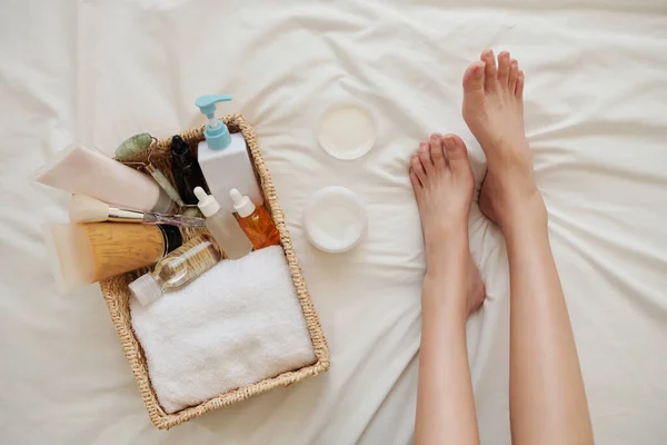 クリーム ローション オイルなどのスキンケア製品のバスケットの横にあるベッドの上の若い女性の足 — ストック写真