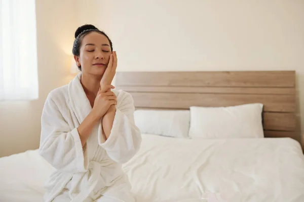 Smiling young woman in bathrobe enjoying touching her soft tender skin after shower
