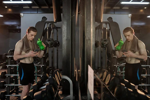 在健身房锻炼期间 健身运动员喝水 并在智能手机上查看社交媒体 — 图库照片