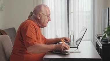 Gözlüklü yaşlı adamın not alıp, bardaktan su içtiği ve evdeki oturma odasındaki masada dizüstü bilgisayarda çalıştığı yan görüntüler.