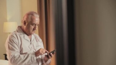 Açıklama, evde pijamalı, yatakta oturan, mesaj yazan ya da cep telefonundaki numarayı arayan son sınıf öğrencisinin aşağı eğilmiş görüntüsü.