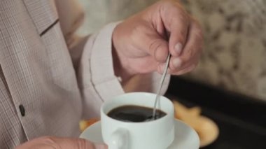 Tanımlanamayan yaşlı bir adamın elinde fincan fincan tutarken kaşıkla kahveyi karıştırırken mutfakta içkiyi hazırlarken çekilmiş görüntüler.
