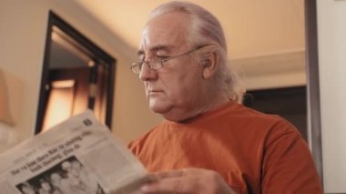 Alçak açılı göğüs üstü, gözlüklü yaşlı adamın evde gazete okuduğu fotoğraf.