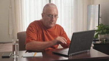 Gözlüklü üst düzey bir adamın masada oturup dizüstü bilgisayarda çalışırken ve evde deftere not alırken orta boy bir fotoğrafı.